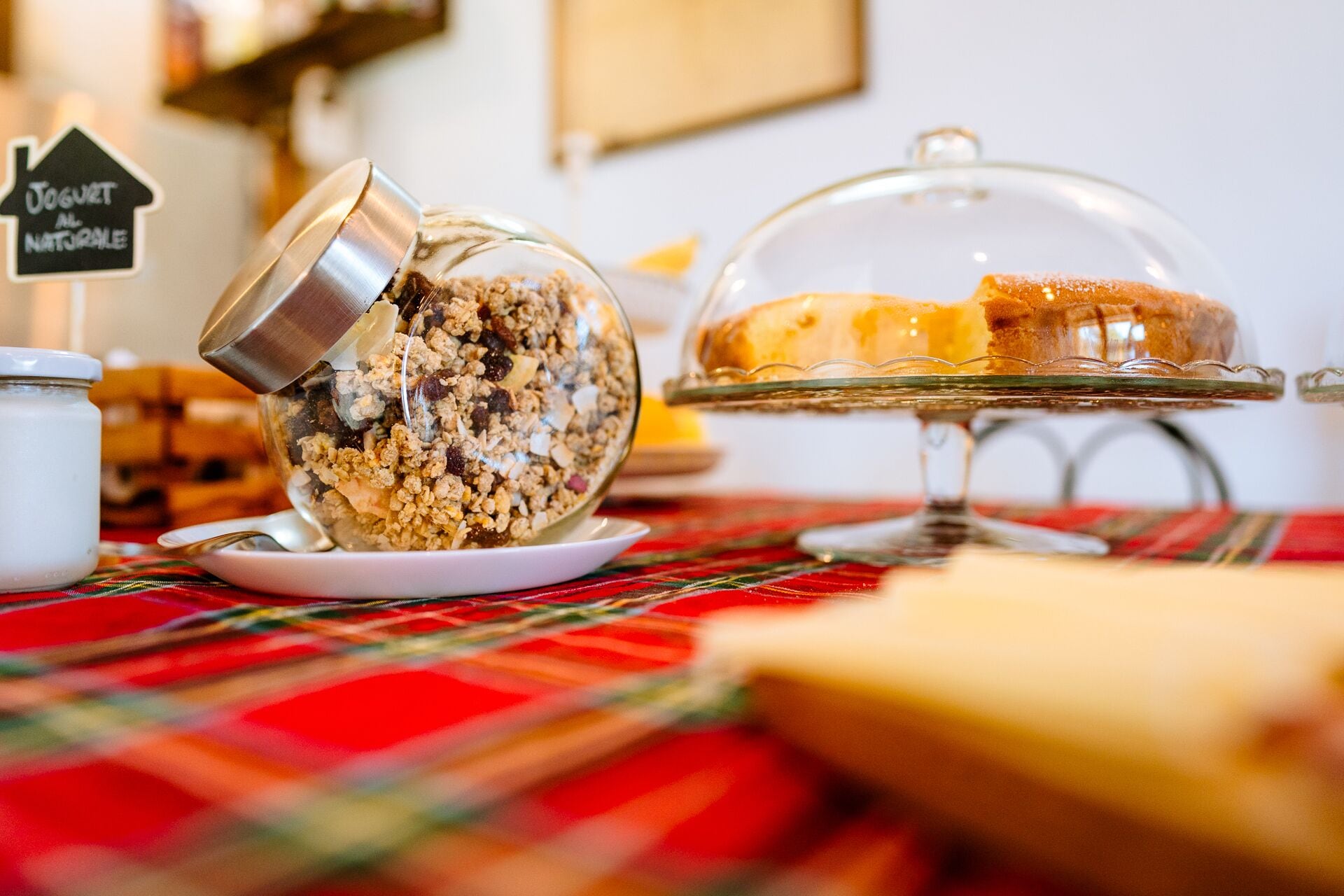 Une table mise pour le petit-déjeuner, avec un bol en verre rempli de céréales, un plat en verre couvert contenant une part de gâteau et un pot de lait, le tout posé sur une nappe à carreaux.