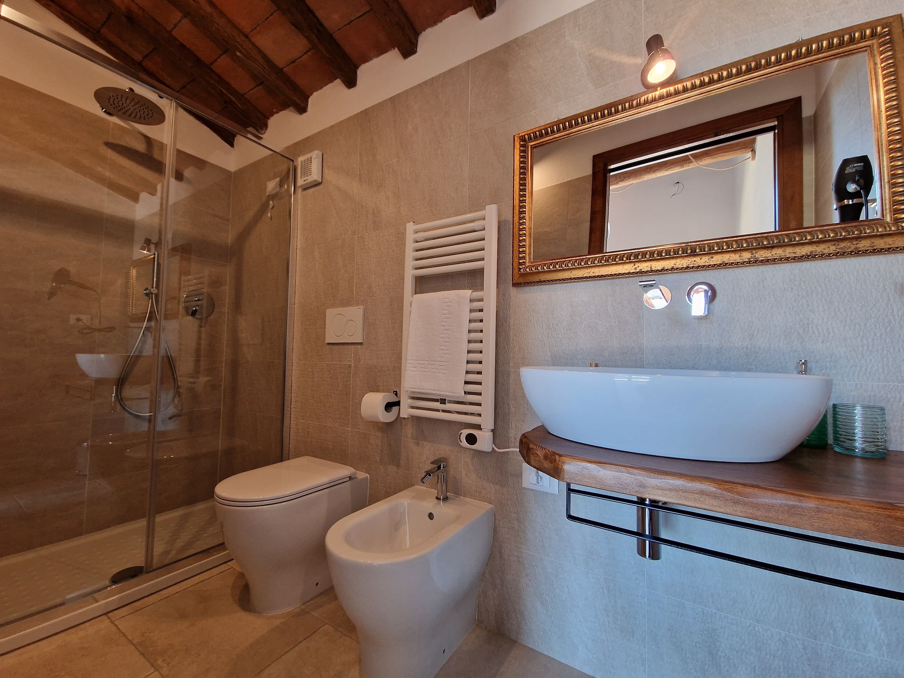 Une salle de bain dotée d’une douche à parois de verre, de toilettes, d’un bidet, d’un lavabo avec un plan de travail en bois et d’un miroir avec un cadre doré.