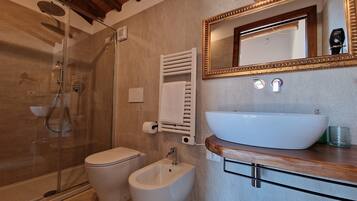Une salle de bain dotée d’une douche à parois de verre, de toilettes, d’un bidet, d’un lavabo avec un plan de travail en bois et d’un miroir avec un cadre doré.