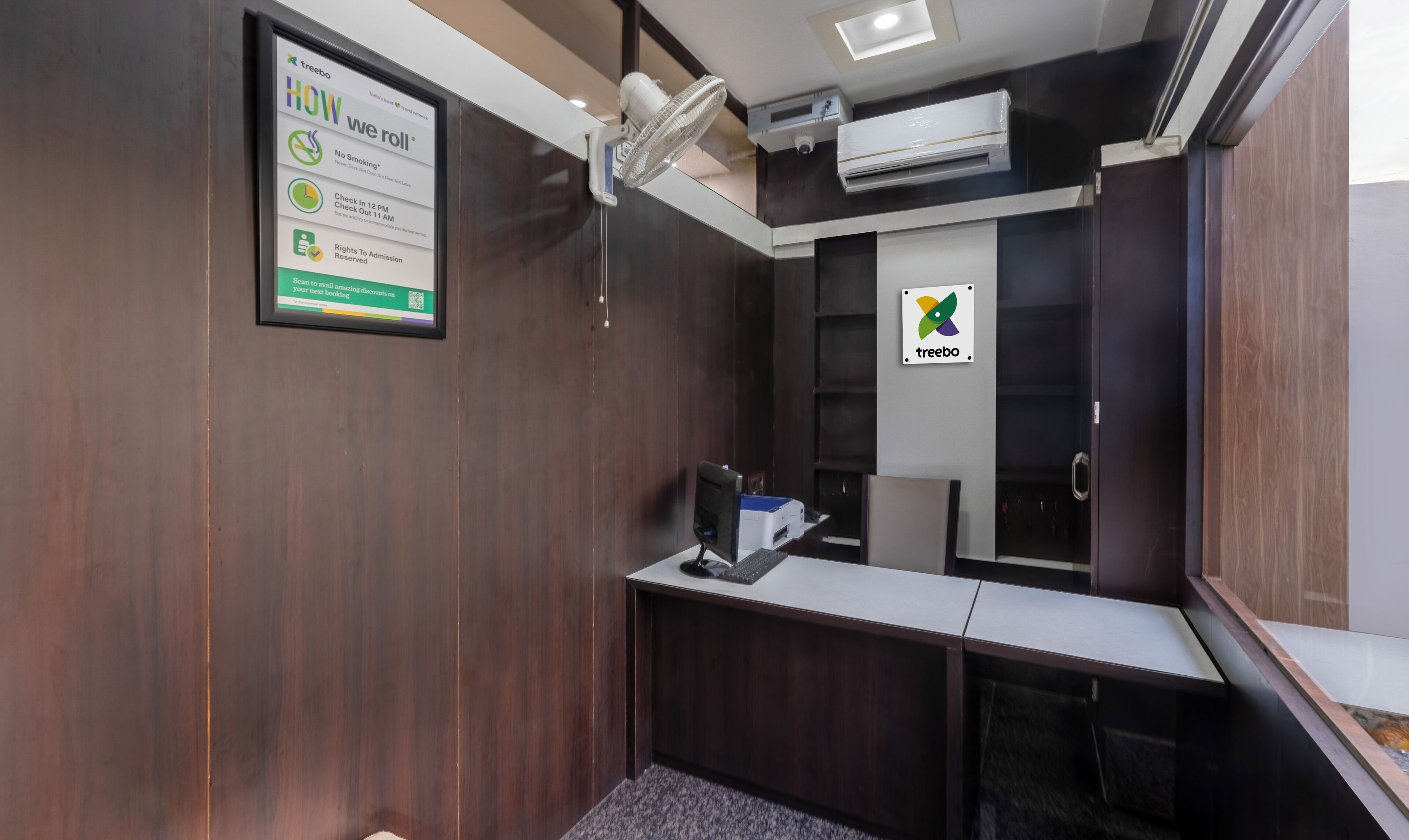 A compact, modern room with a desk, a chair, and a large mirror. There is a framed sign on the wall with information and a logo.
