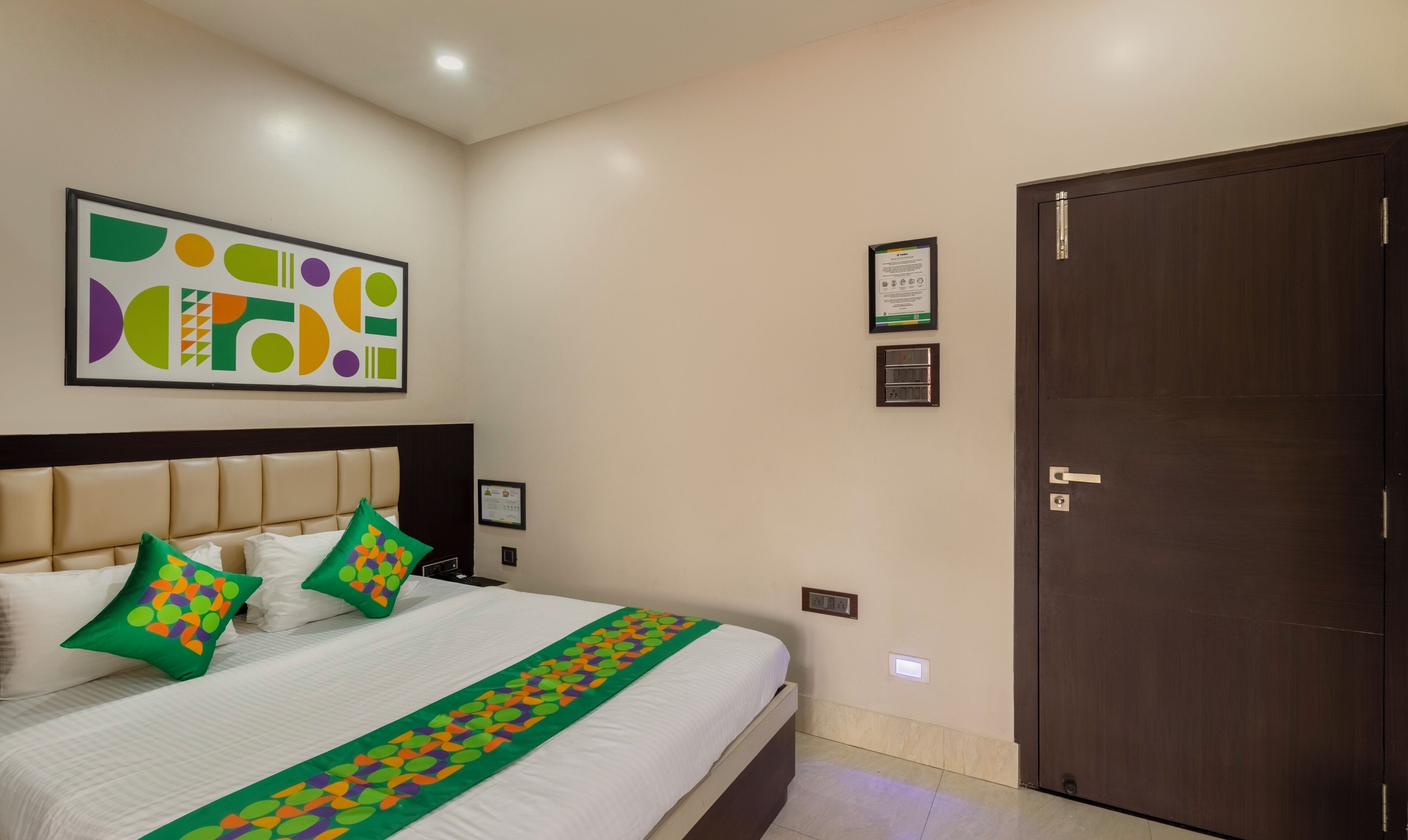 A hotel room with a large bed, a headboard, a door, and a wall with a framed abstract artwork.