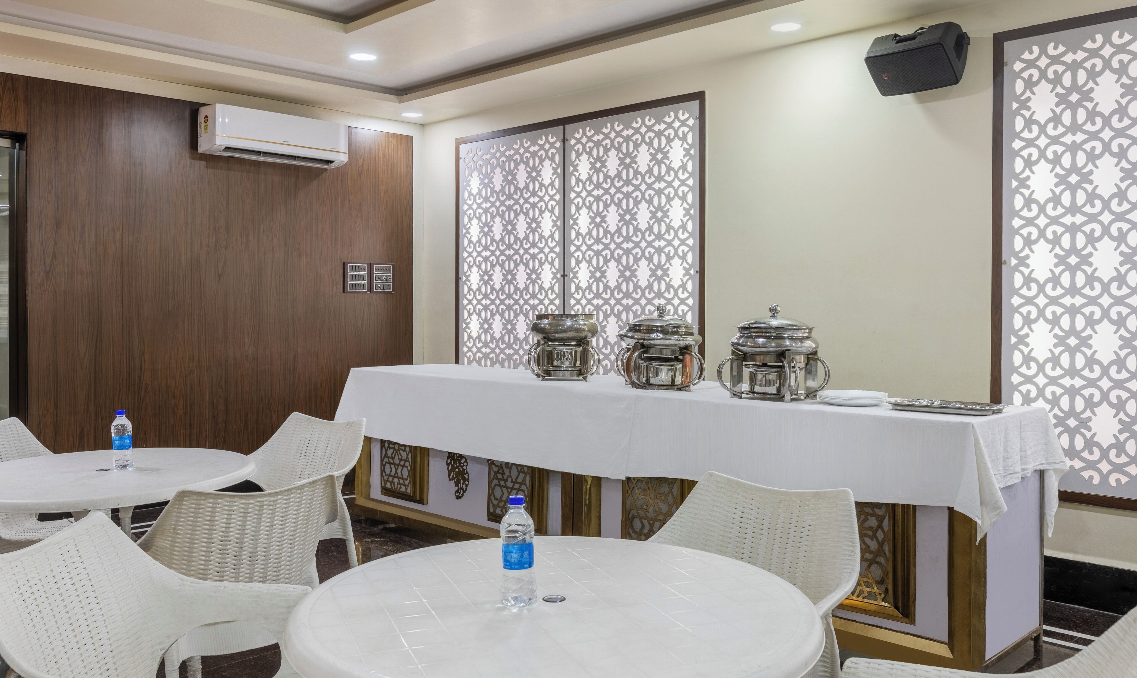 A hotel room with a dining area featuring round tables and white chairs, a decorative partition, and a wall-mounted air conditioner.