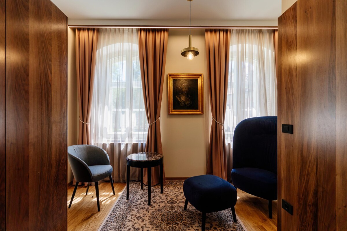 Ein Zimmer mit Holzwandverkleidung, einem dunkelblauen Sessel, einem kleinen Stuhl, einem kleinen Tisch, einem gemusterten Teppich und einem Bild an der Wand.