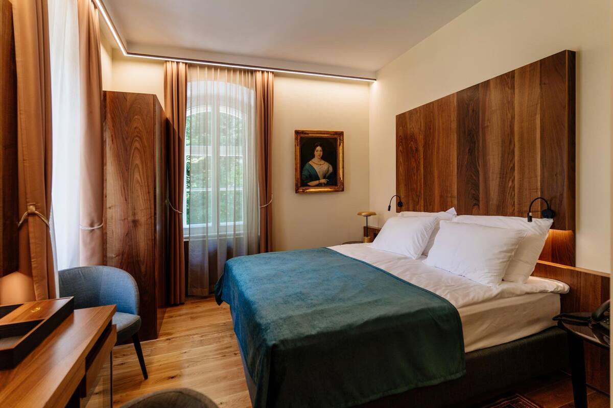 A hotel room with a bed, a desk, a chair, and a painting on the wall.