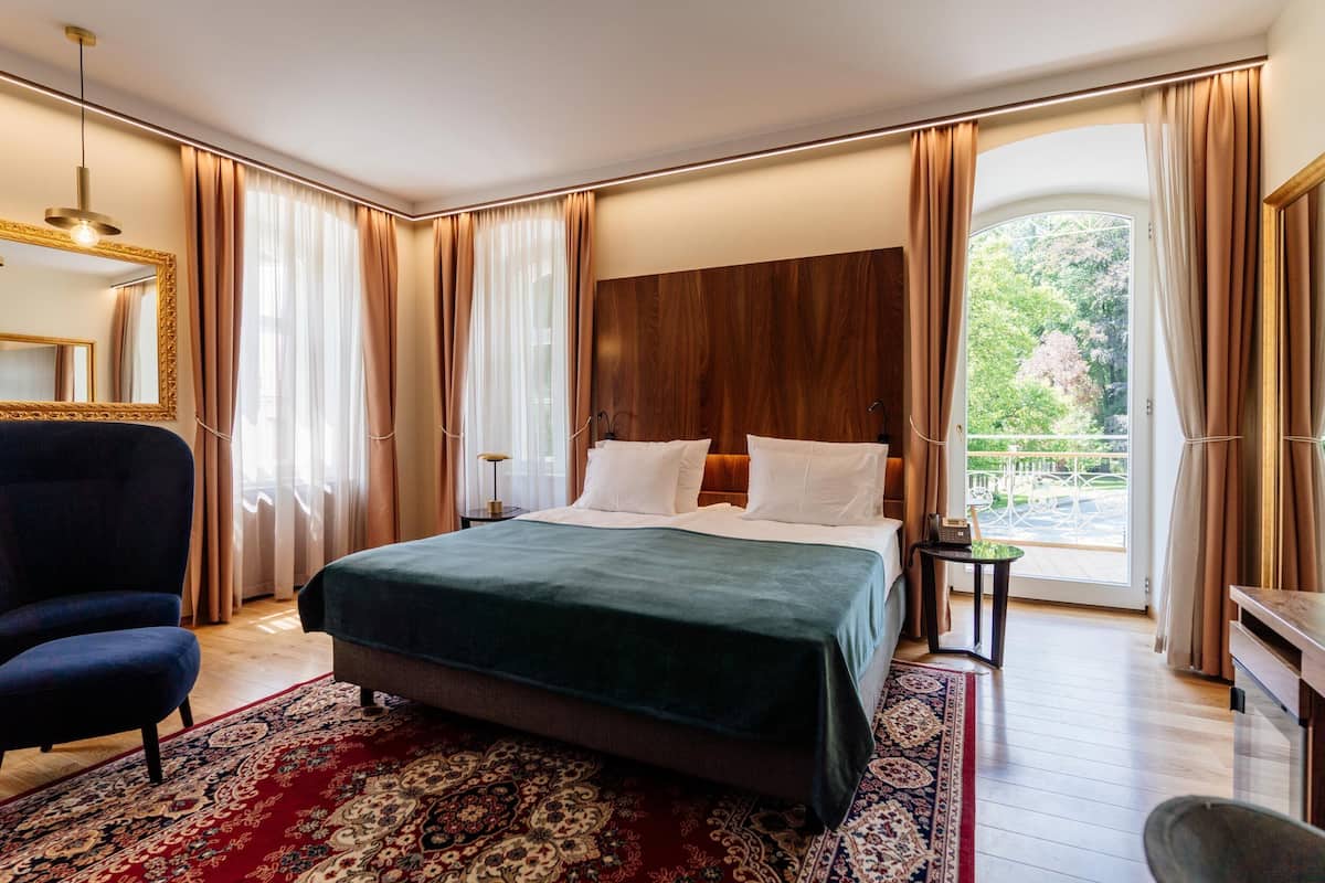 A spacious bedroom with a large bed, a sitting area with a chair, a small table, and a view of the outdoors through a large window.