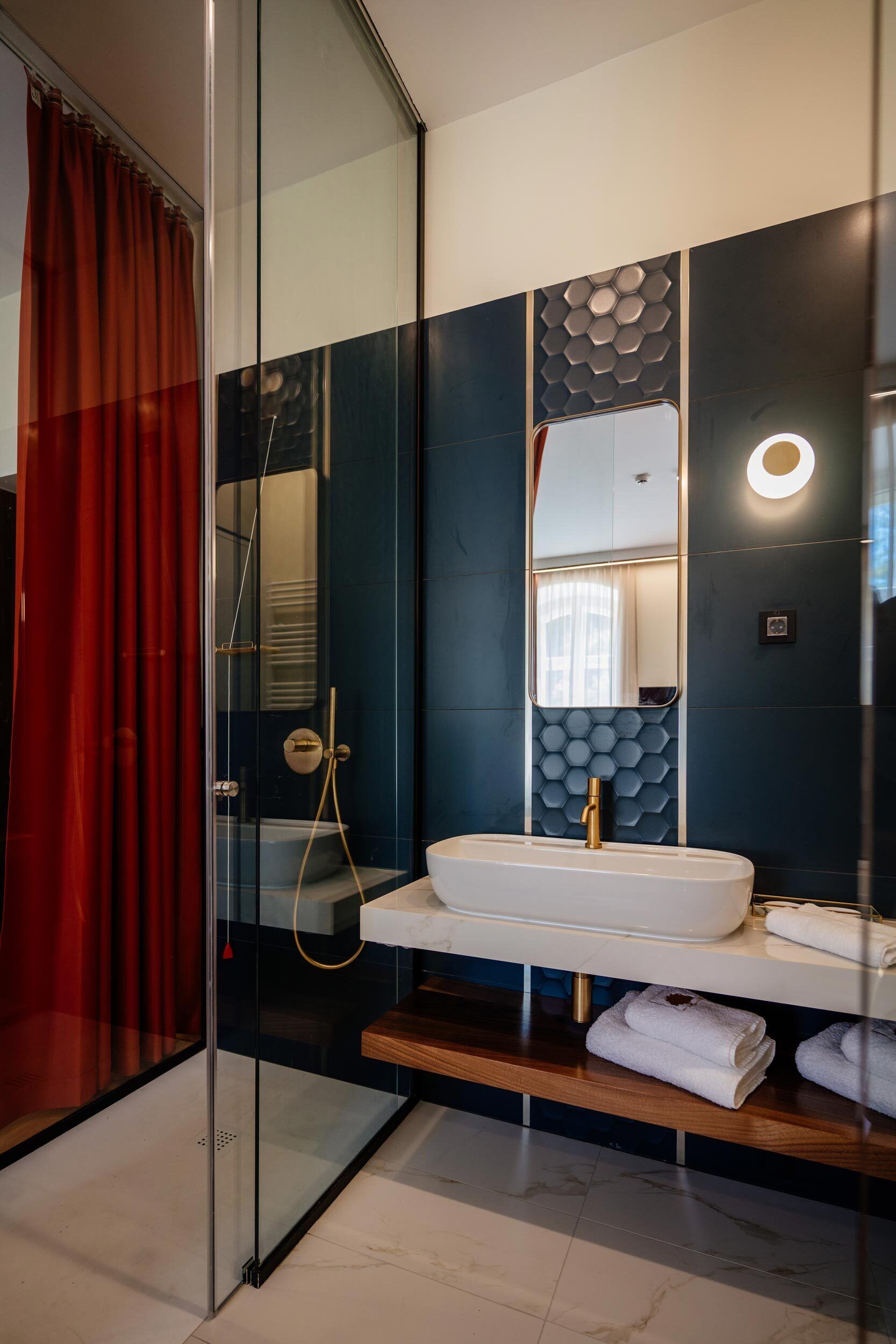 Un baño moderno con cabina de ducha de vidrio, un lavamanos blanco con grifo dorado y un espejo con diseño hexagonal en la pared.
