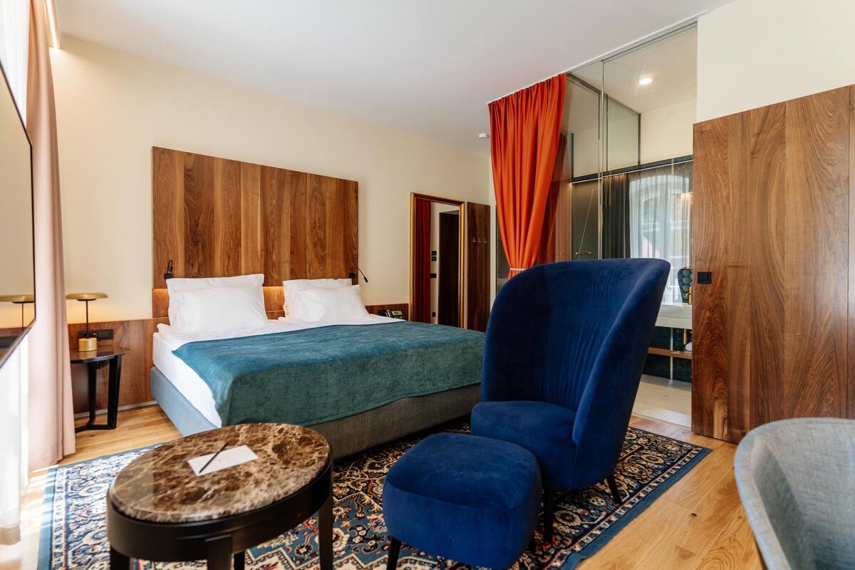 A hotel room with a large bed, a blue armchair, a small round table, and a patterned rug.