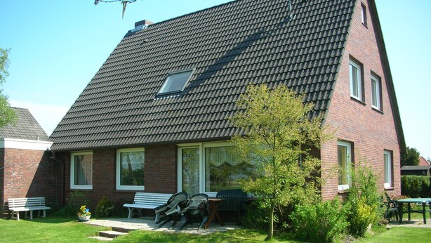 A two-story house with a sloped roof, a small window, and a garden with benches and a tree. - Beautiful fine apartment with garden, meadow, view of water and land (Dornummersiel/Dornum)
