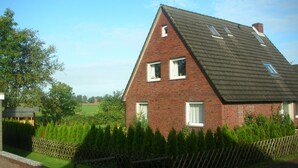 A two-story brick house with a sloped roof, multiple windows, and a well-maintained garden with a wooden fence. - Beautiful fine apartment with garden, meadow, view of water and land (Dornummersiel/Dornum)