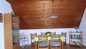 A dining area with a wooden ceiling, a table with chairs, a bookshelf, and a refrigerator. - Beautiful fine apartment with garden, meadow, view of water and land (Dornummersiel/Dornum)