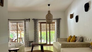 Un salón con sofá, mesa de centro y lámpara colgante. Se observan cortinas estampadas y vistas de vegetación al exterior.