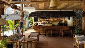 Una zona de bar con barra de madera, techo de paja, mobiliario de madera y una variedad de botellas en estantes.