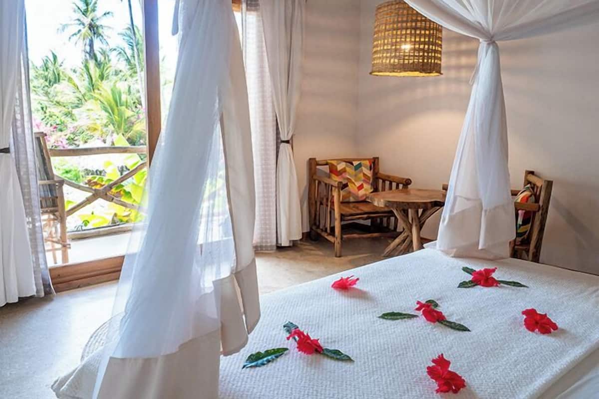Habitació amb un llit, una taula de fusta i cadires. Hi ha una finestra amb cortines i vistes a la vegetació exterior.