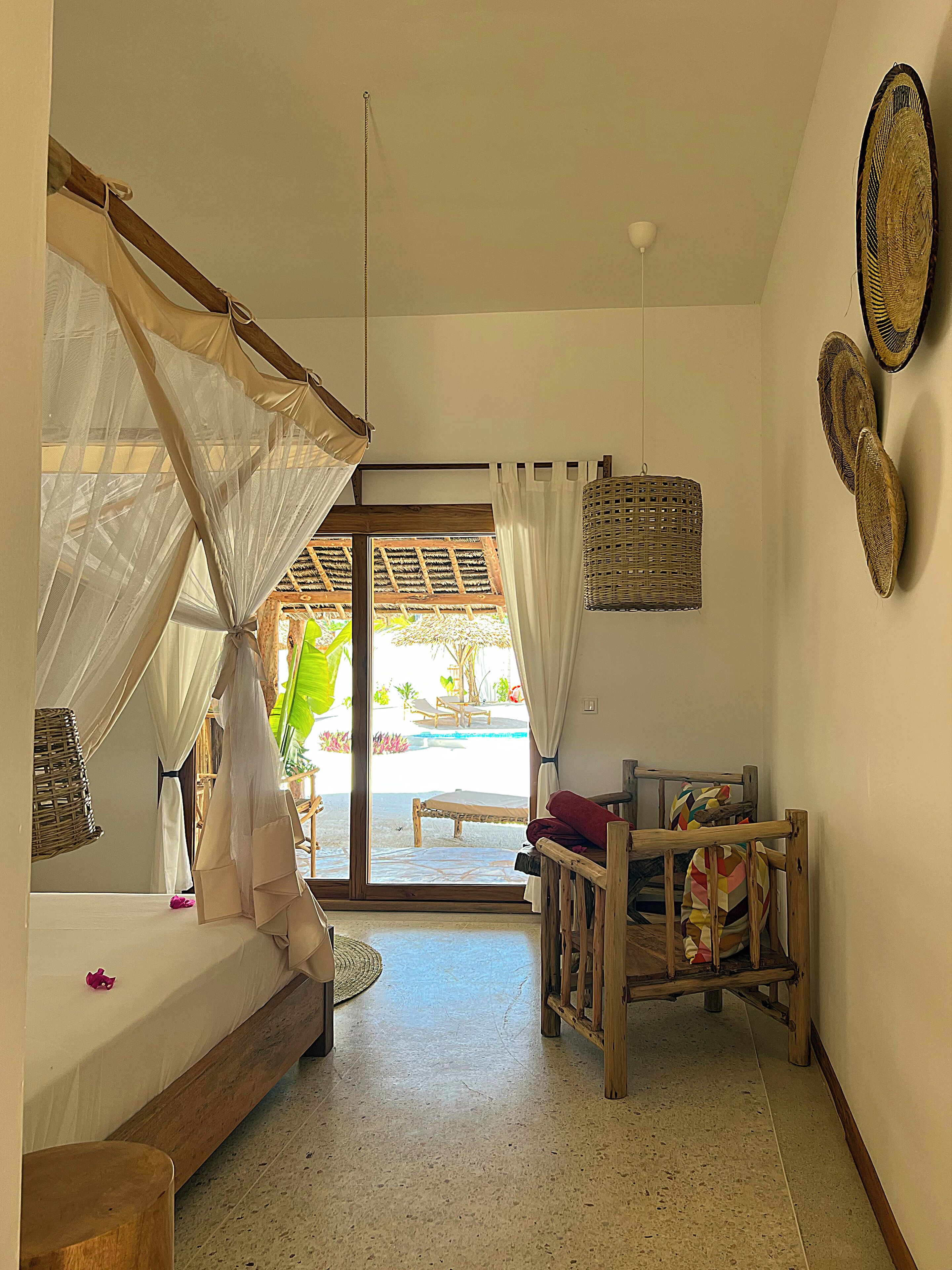 Una habitación con una cama, un banco de madera, una cesta de mimbre y vistas a un área exterior con piscina y sillas.