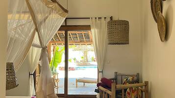 Una habitación con una cama, un banco de madera, una cesta de mimbre y vistas a un área exterior con piscina y sillas.