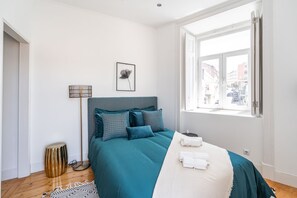 Een keurig opgemaakt slaapkamer met een bed, nachtkastje, lamp en een raam met uitzicht op gebouwen.