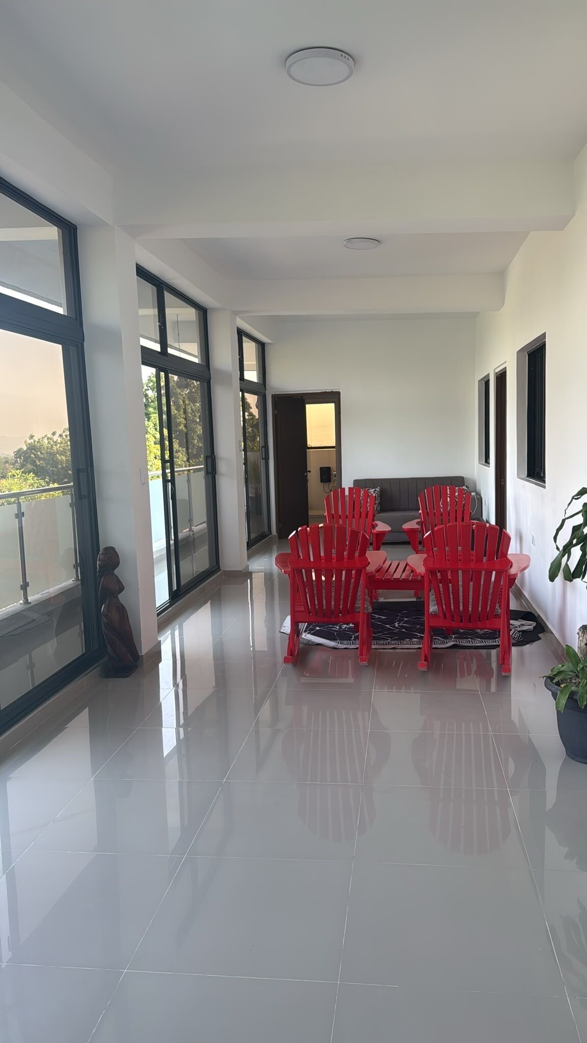 A spacious room with a modern design, featuring a dining area with red chairs and a table, large windows, and a view of the outdoors.