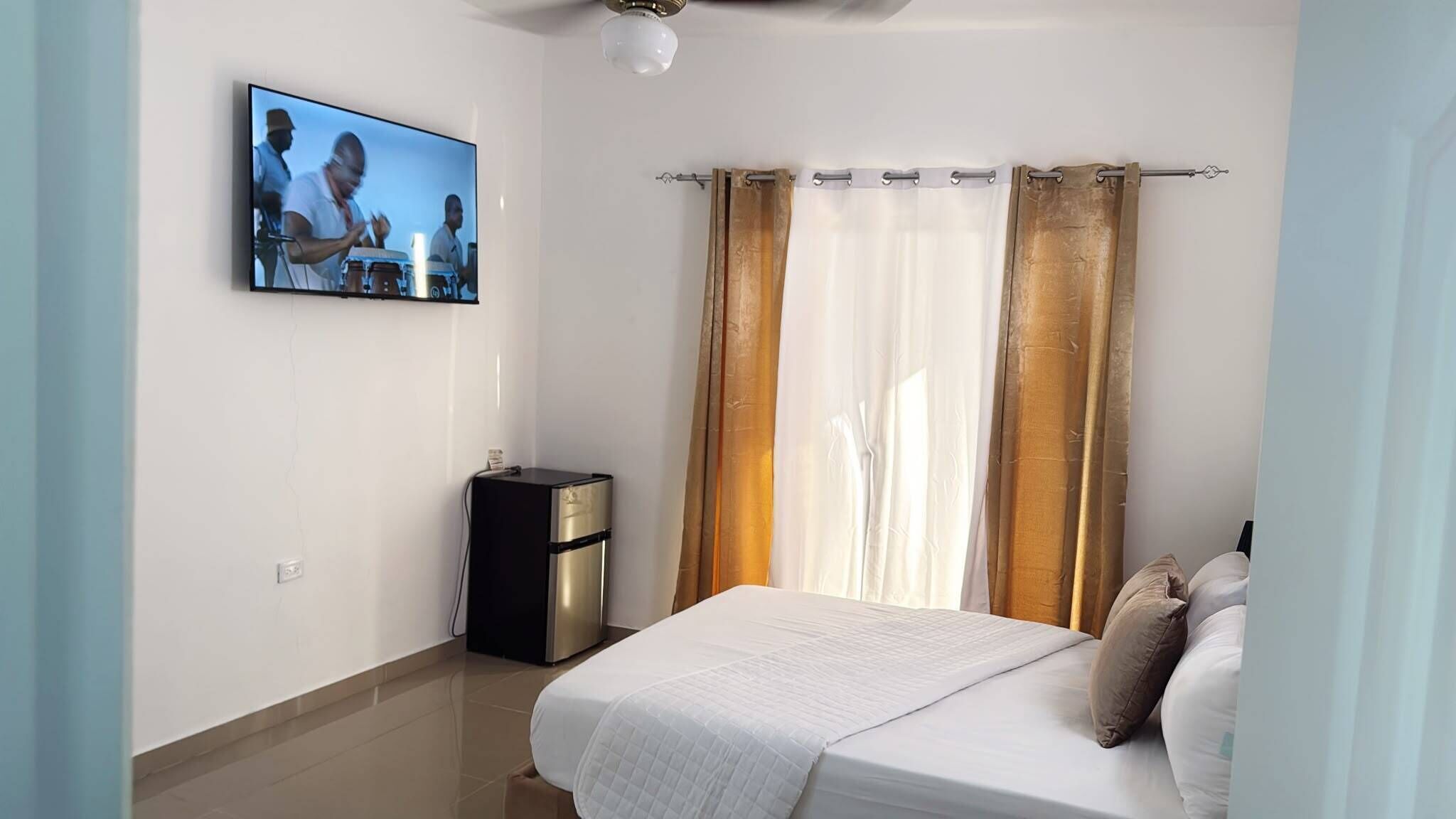 A bedroom with a bed, a TV mounted on the wall, a bedside table, and curtains.