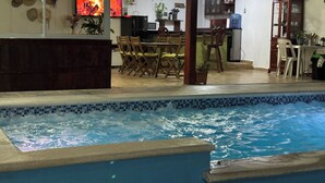 A swimming pool with a tiled edge, a dining area with wooden chairs and a table, and a kitchen area with cabinets and a water dispenser.
