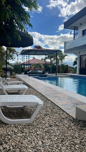 Una piscina con sillones de descanso, un área de estar cubierta y un edificio moderno al fondo.
