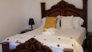 A neatly made bed with a wooden headboard, white bedding, and a single yellow pillow. There are three wall-mounted light switches and a small table with a lamp beside the bed.