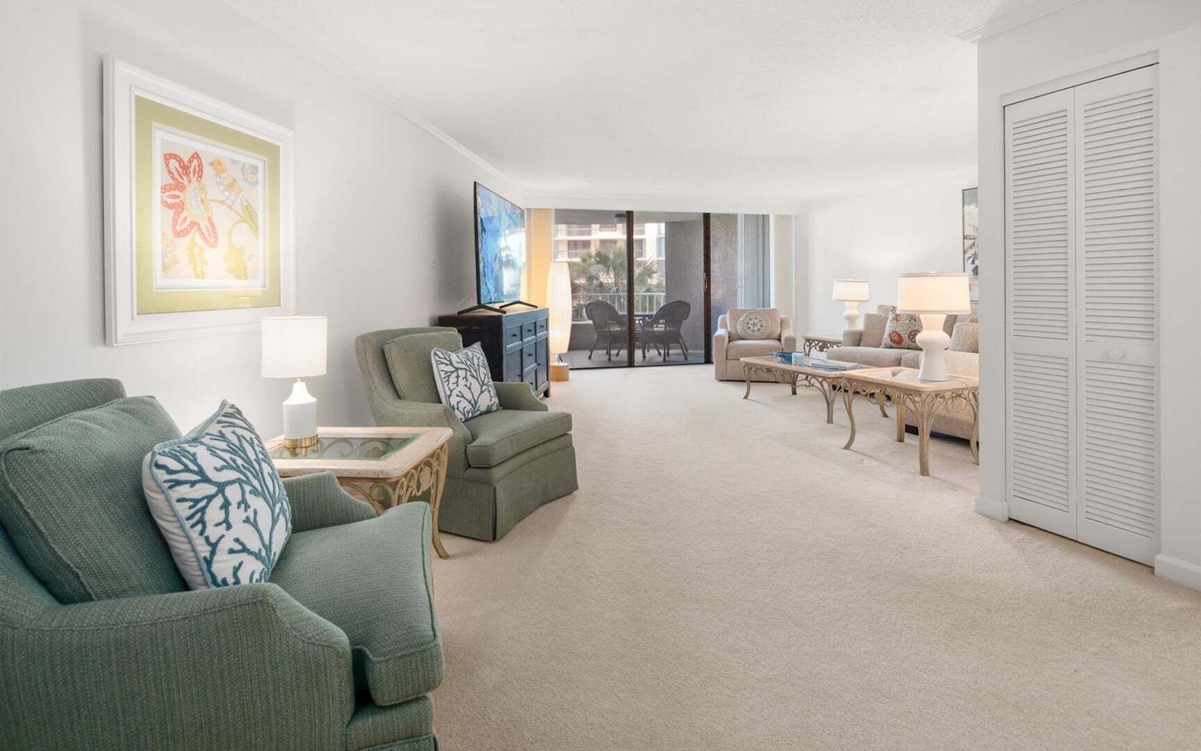 A living room with a green armchair, a sofa, a coffee table, and a TV. There is a door with a white closet and a window with blinds.