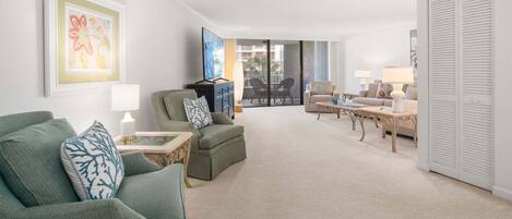 A living room with a green armchair, a sofa, a coffee table, and a TV. There is a door with a white closet and a window with blinds.