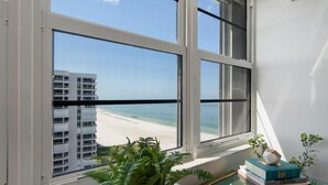 Una habitación con un ventanal grande que da a una playa y un edificio. Hay una planta en maceta y una pila de libros sobre una mesa de vidrio.