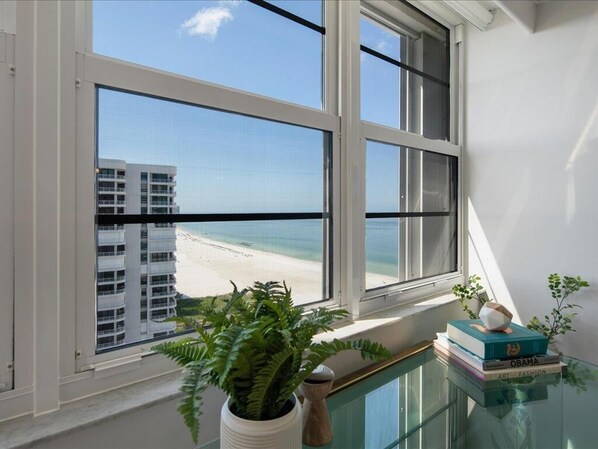A room with a large window overlooking a beach and a building. There is a potted plant and a stack of books on a glass table. - Beachfront 3BR/3BA condo with pool, Wi-Fi, and resort-style comfort. (Marco Island)