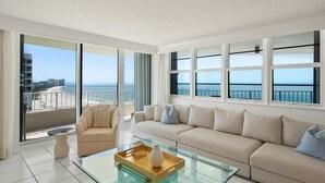 Una sala de estar moderna con una mesa de centro de vidrio, sofá color beige y amplios ventanales con vista a la playa y al océano.