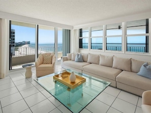A modern living room with a glass coffee table, beige sofa, and large windows offering a view of the beach and ocean.