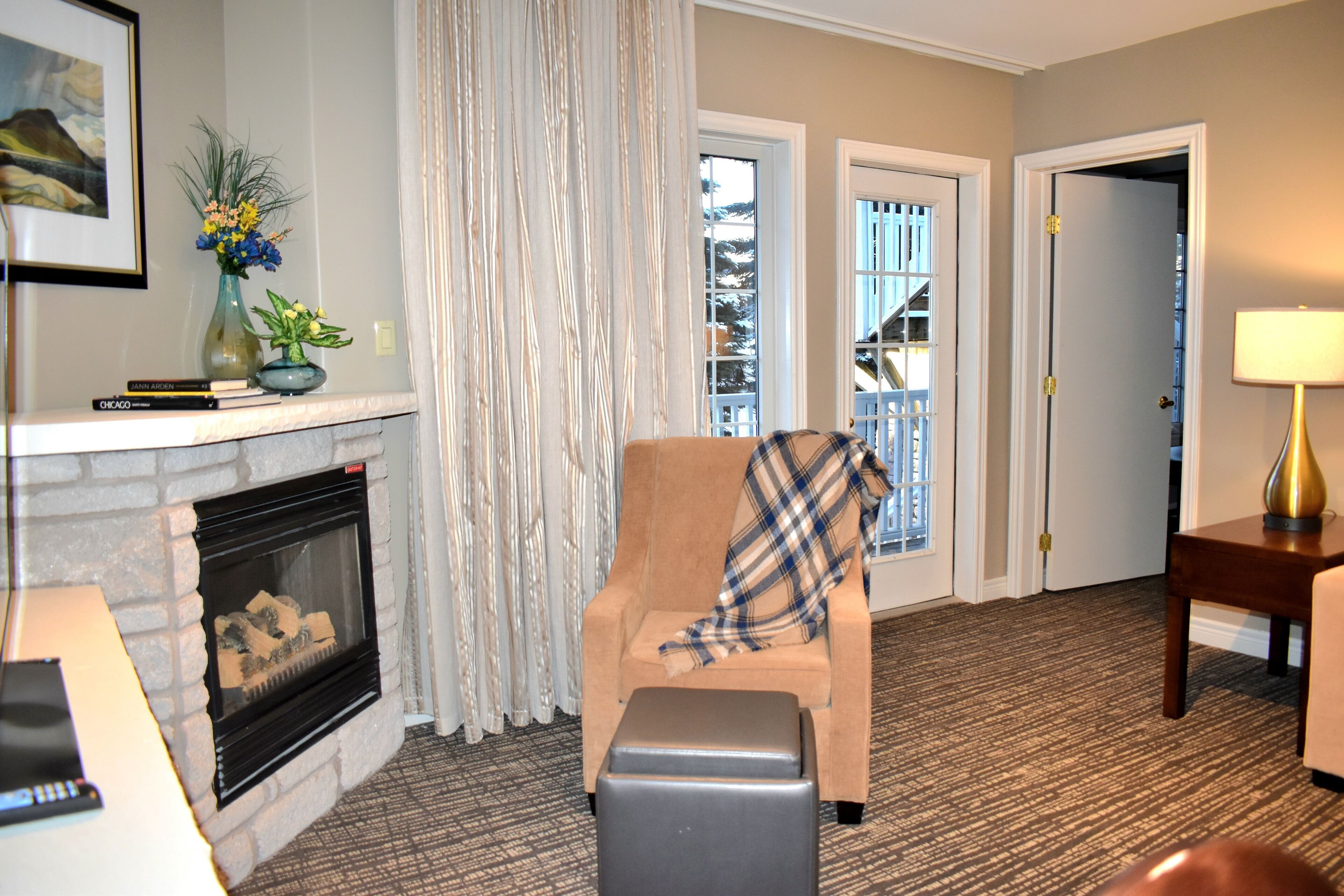 A living room with a fireplace, a plaid chair, a small table with a lamp, and a door leading to a balcony.