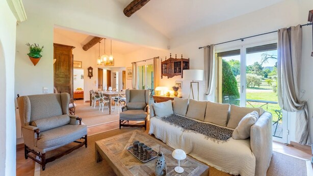 Un salon avec un canapé, des fauteuils et une table basse. On aperçoit un coin repas au fond de la pièce.