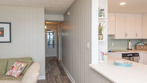 A modern kitchen with white cabinets, a countertop, and a backsplash. There is a dining area with a green couch and a colorful pillow. - Newly renovated 2 bedroom, 2 bath condo with beautiful views of the ocean! (Ocean City)