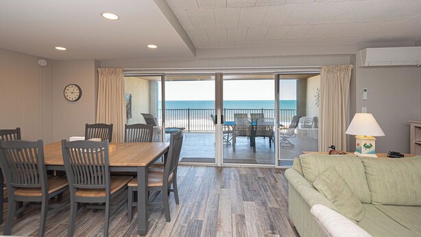 A room with a wooden dining table, chairs, a sofa, and a view of the ocean through large sliding glass doors. - Newly renovated 2 bedroom, 2 bath condo with beautiful views of the ocean! (Ocean City)