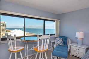 A room with a large window overlooking the sea, a small round table, two white chairs, a blue sofa, and a white nightstand with a lamp. - Spacious, comfortable 2 bedroom oceanfront condo with free WiFi, an outdoor pool, and an enclosed balcony with a great ocean view located midtown only steps to the beach! (Ocean City)