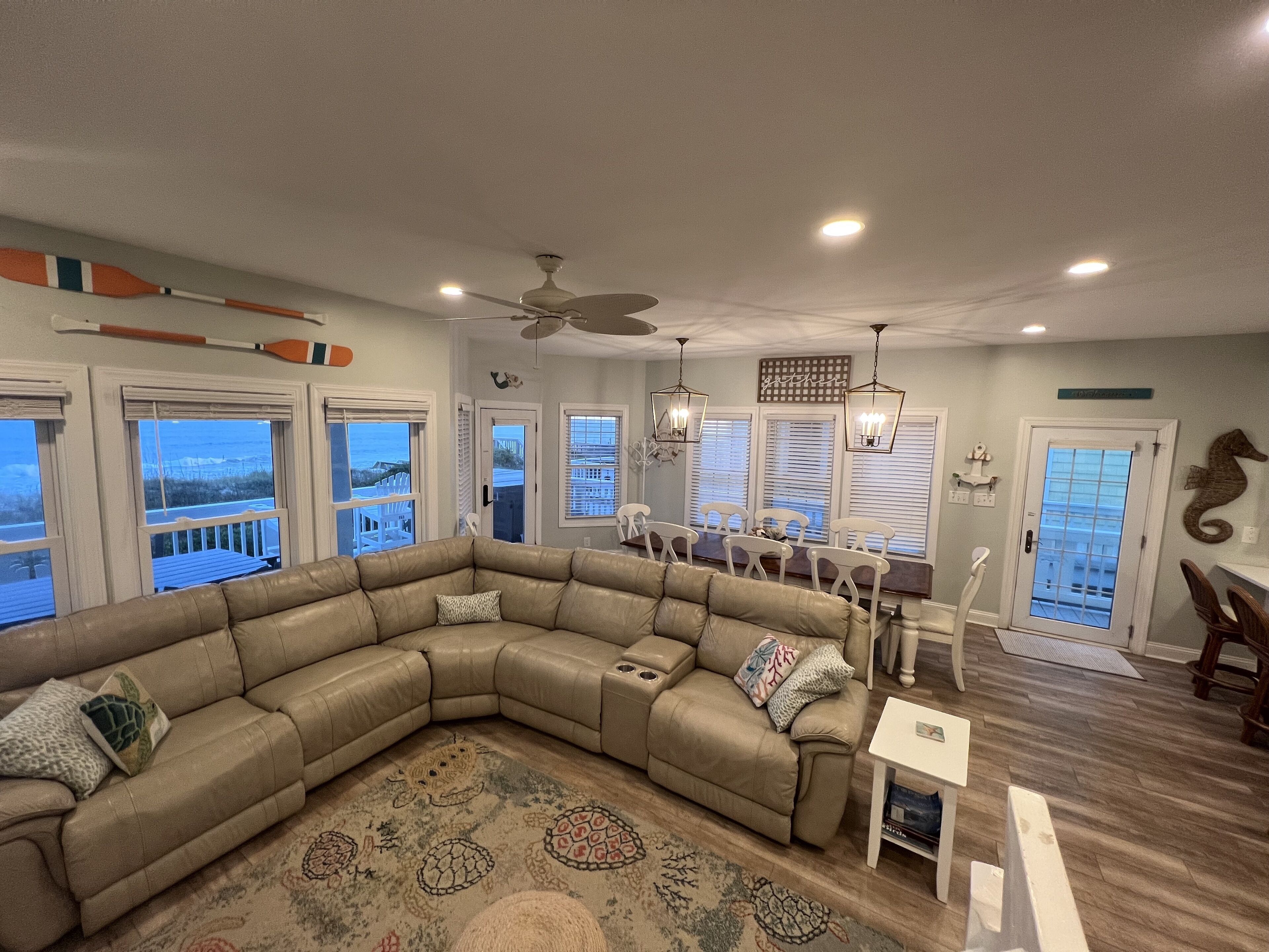 A spacious living room with a large L-shaped sectional sofa, a dining area with a table and chairs, and a view of the ocean through large windows.