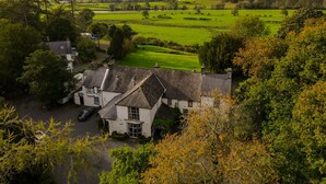 A large white house with a steep roof, surrounded by lush greenery and a view of rolling hills. - Plas Dolguog (Machynlleth)