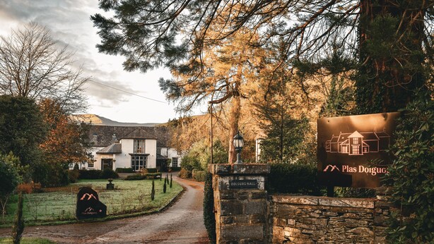 A rural road leading to a white building with a stone wall entrance, a sign for 'Plas Doigog', and a mountain silhouette. - Plas Dolguog (Machynlleth)