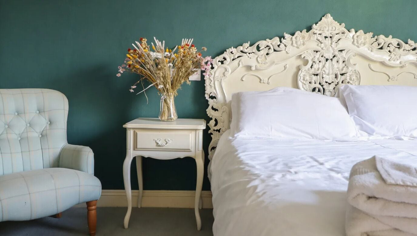 A bedroom with a white upholstered armchair, a bedside table with a vase of dried flowers, and a white ornate headboard.