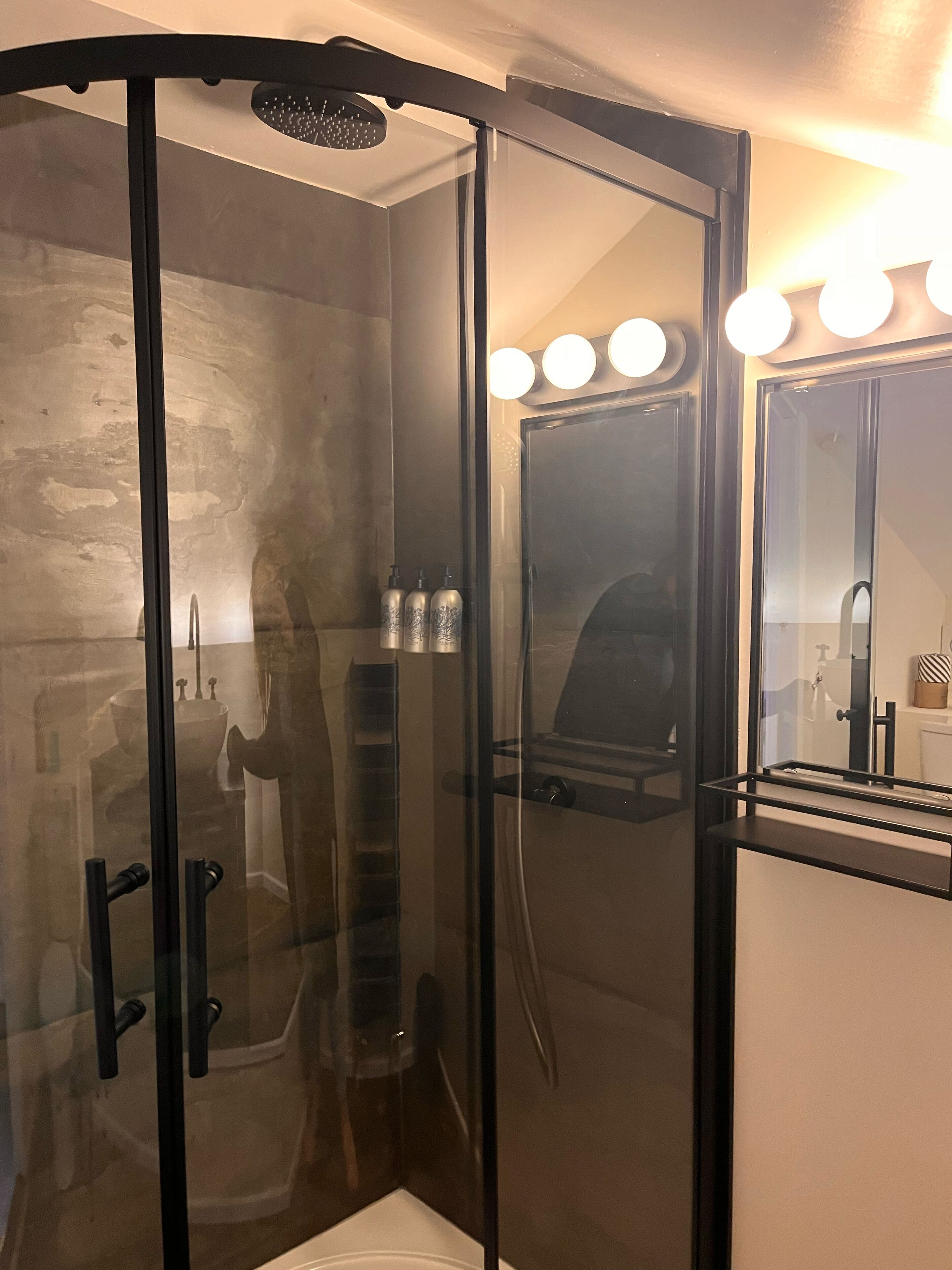 A glass-enclosed shower with a black frame, a sink with a mirror, and three bottles of shampoo.