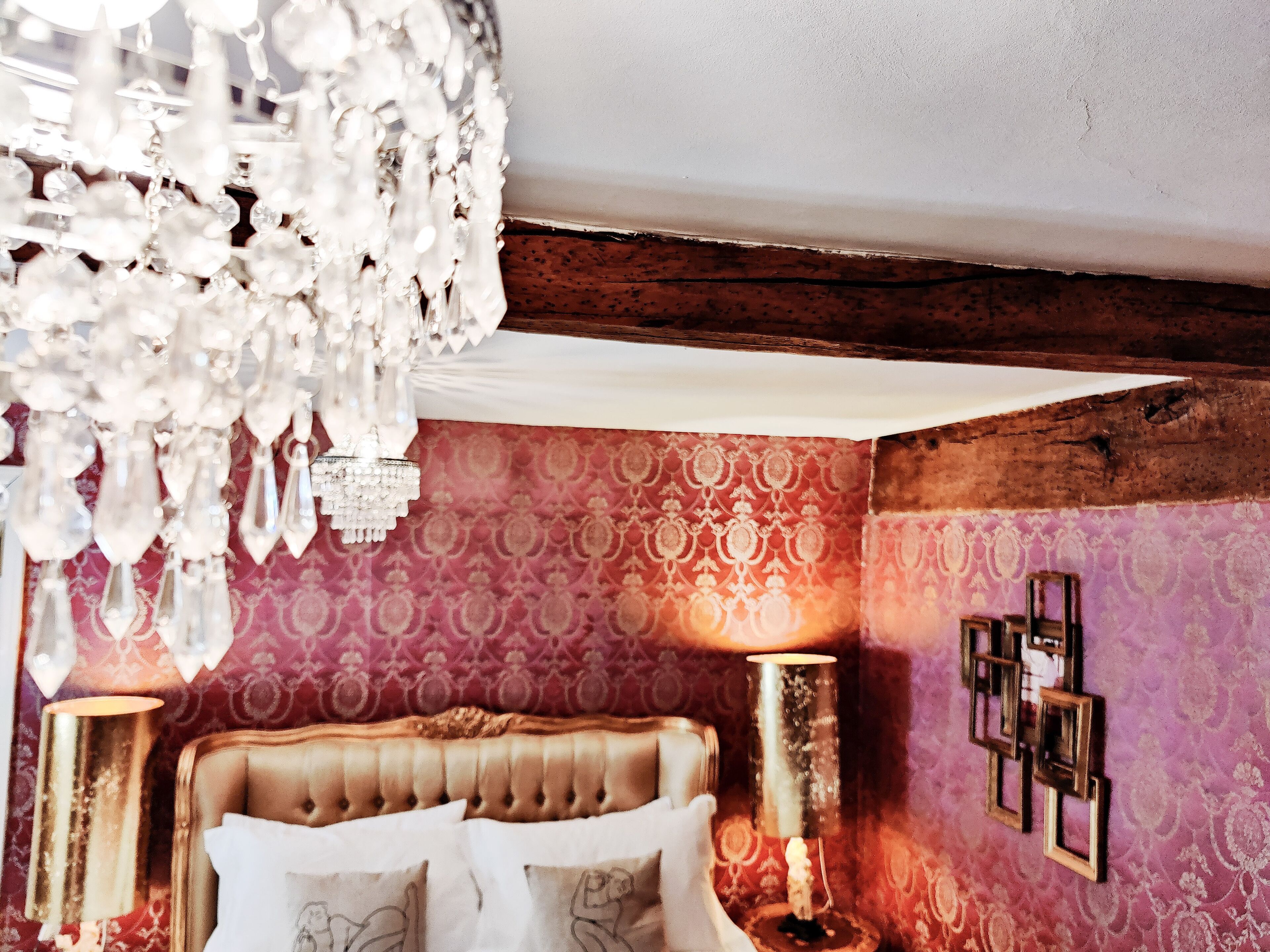 A bedroom with a tufted headboard, a chandelier, patterned wallpaper, and bedside lamps.
