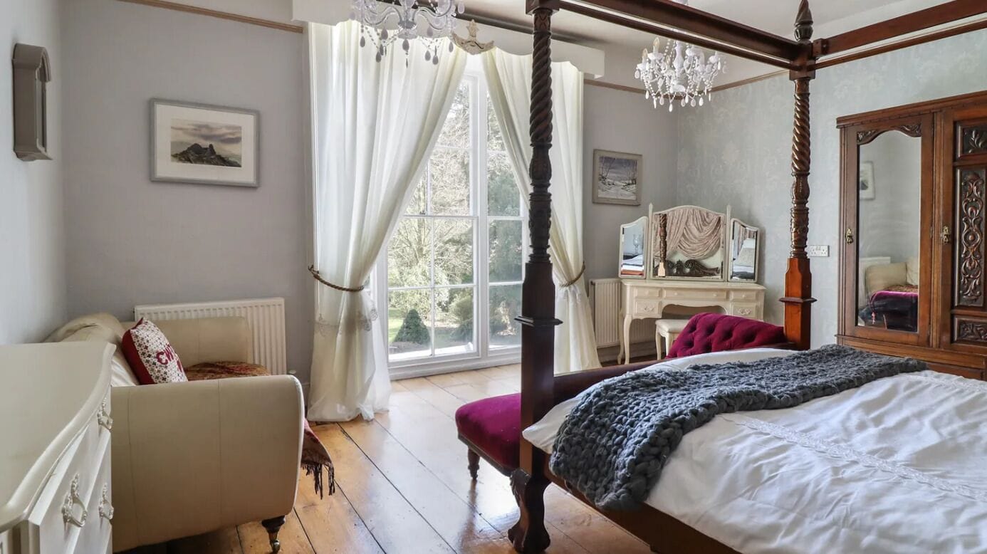 A four-poster bed with a canopy, a wooden desk with a mirror, and a view of the outdoors through a large window.
