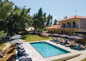 A pool area with lounge chairs and umbrellas, a clear blue pool, and a building with a red-tiled roof. - Pefkon Suites (Kassandra)
