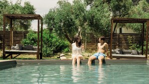 A pool area with two people sitting on the edge, surrounded by wooden pergolas and outdoor furniture.