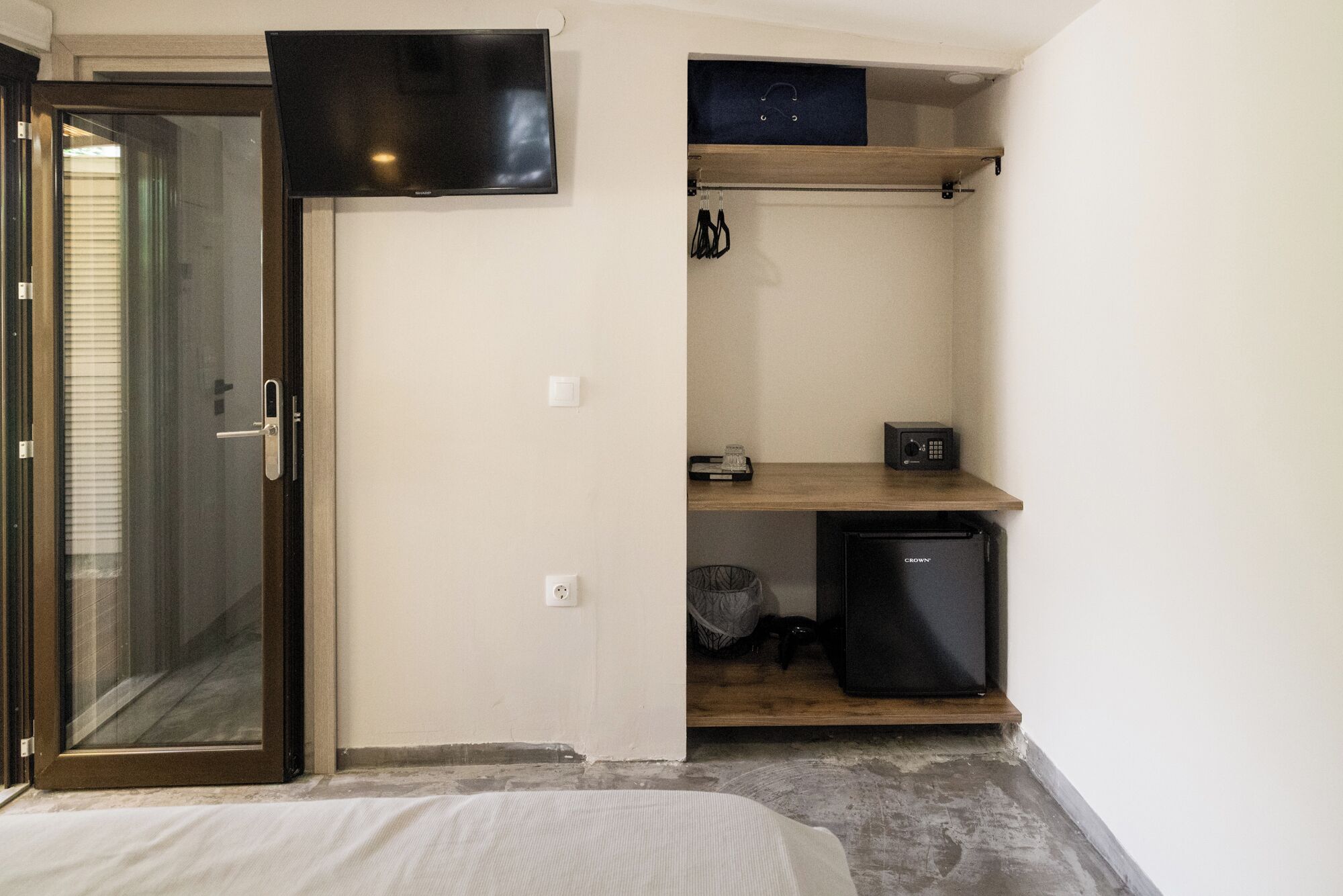 A small, compact room with a bed, a television mounted on the wall, a small refrigerator, and a wooden shelf.