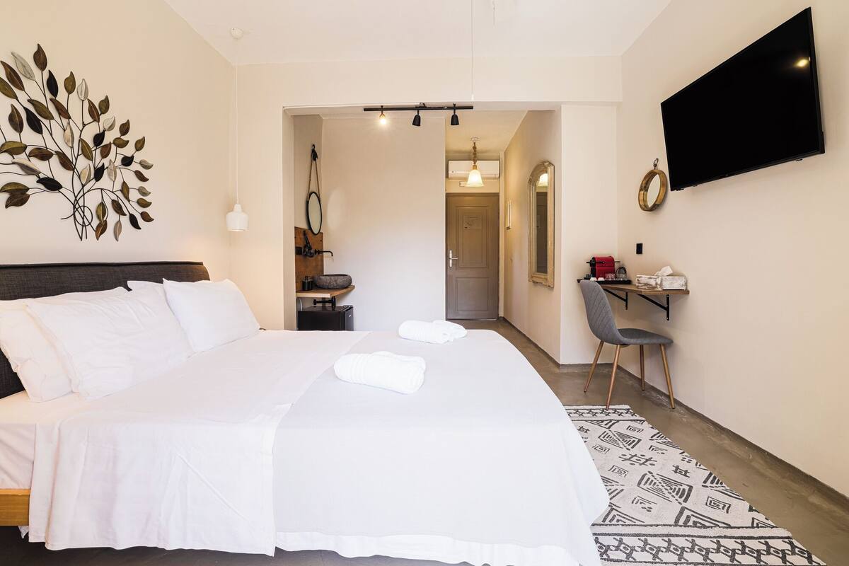 Une chambre d’hôtel avec un grand lit, un bureau avec une chaise, un téléviseur fixé au mur et une salle de bain visible à travers une porte ouverte.