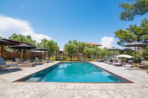 A rectangular swimming pool with lounge chairs and umbrellas around it, set in a sunny outdoor area with trees and a wooden structure in the background. - Pefkon Suites (Kassandra)