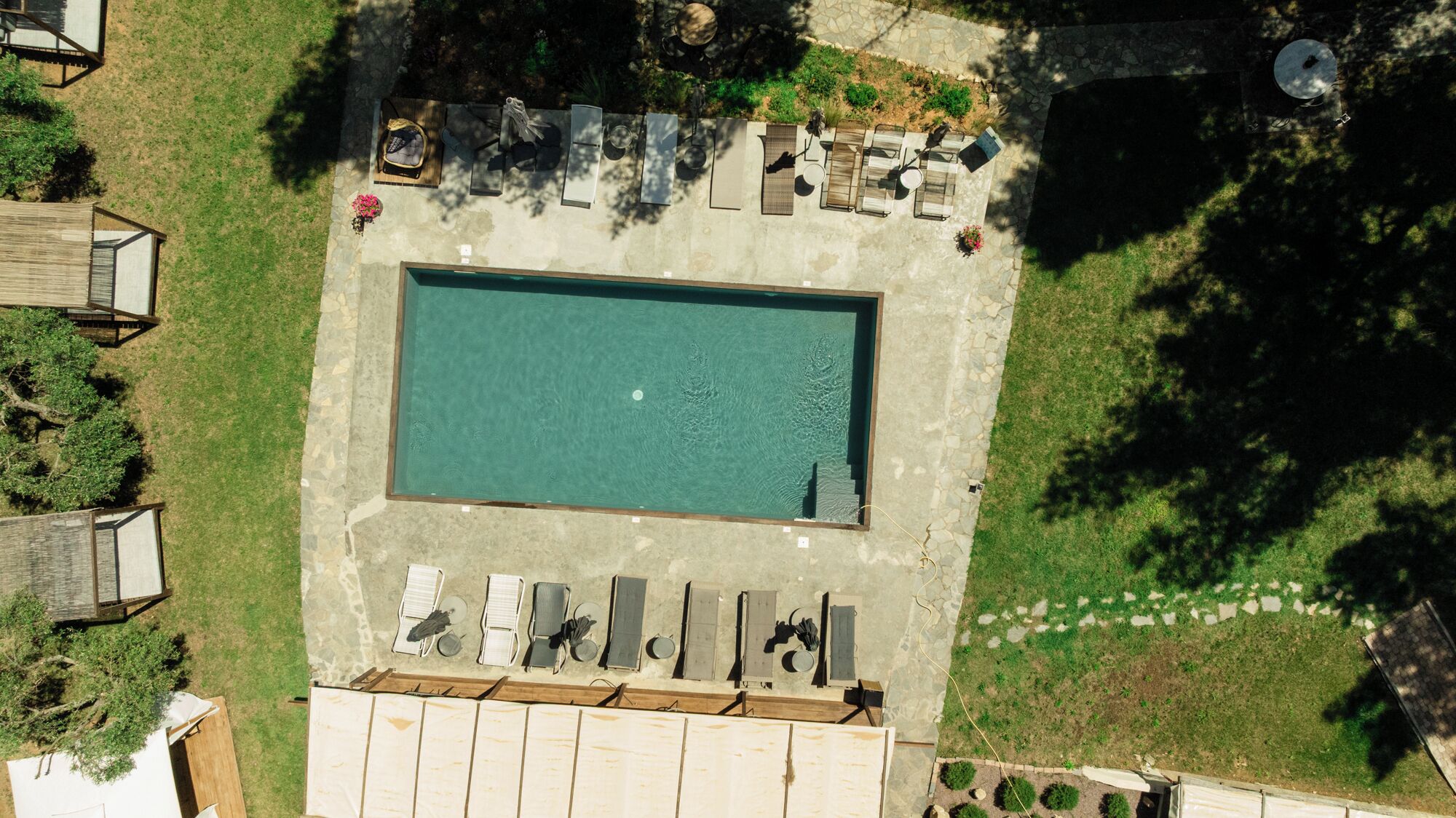An aerial view of a rectangular swimming pool surrounded by lounge chairs and umbrellas, with a stone deck and a small garden area.