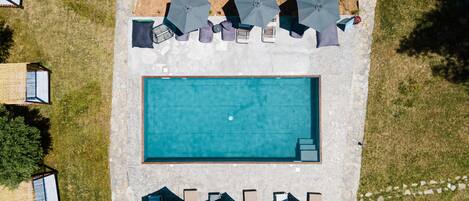 An aerial view of a swimming pool area with lounge chairs and umbrellas, surrounded by stone walls and greenery.